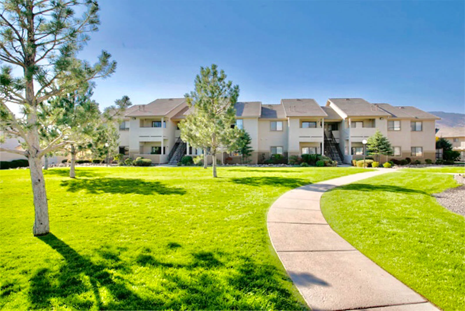 Manicured emerald lawn and a curving walkway in front of a Reno apartment community, framed by pines and high-desert mountains