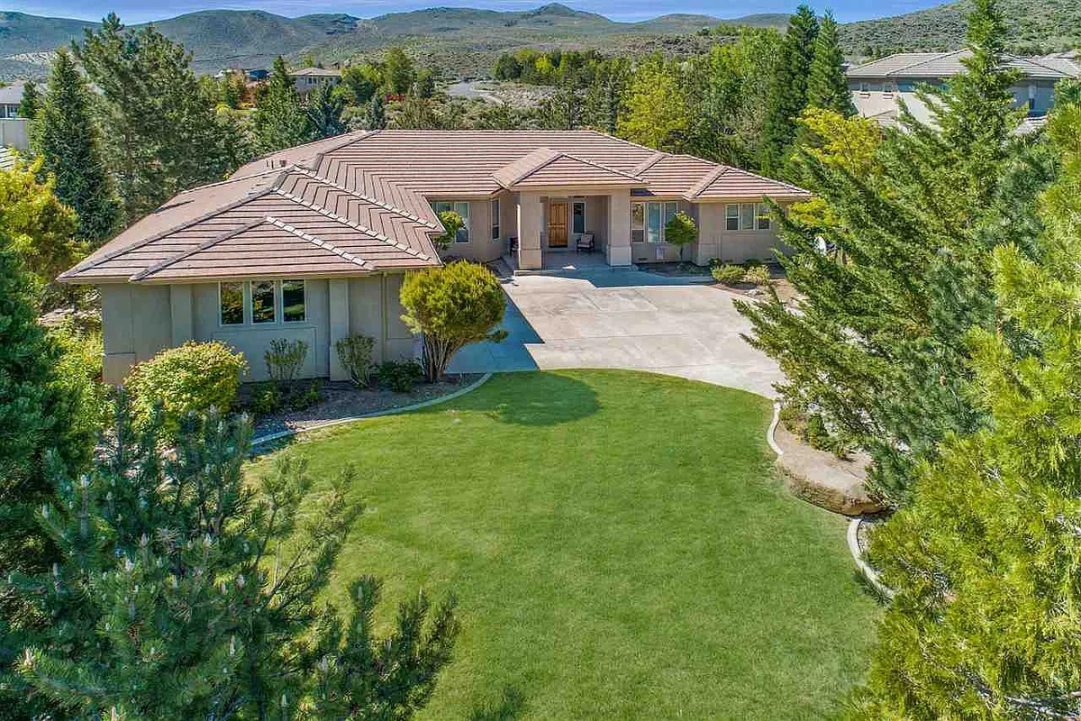 Single-story Reno home with a manicured green lawn, shaped shrubs and a paver driveway, backed by pines and foothills